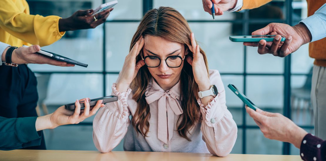 Shot of a stressed out businesswoman surrounded by colleagues needing help. Businesswoman feeling stressed out in a demanding office environment at work. Photo of businesswoman under strain as colleagues request various things from her. Businesswoman feeling overwhelmed by her colleagues in the office. Frustrated businesswoman having a headache while her colleagues are demanding her to work on many things in the office. Stress, anxiety and multitasking businesswoman with headache from workload and laptop deadline in office.