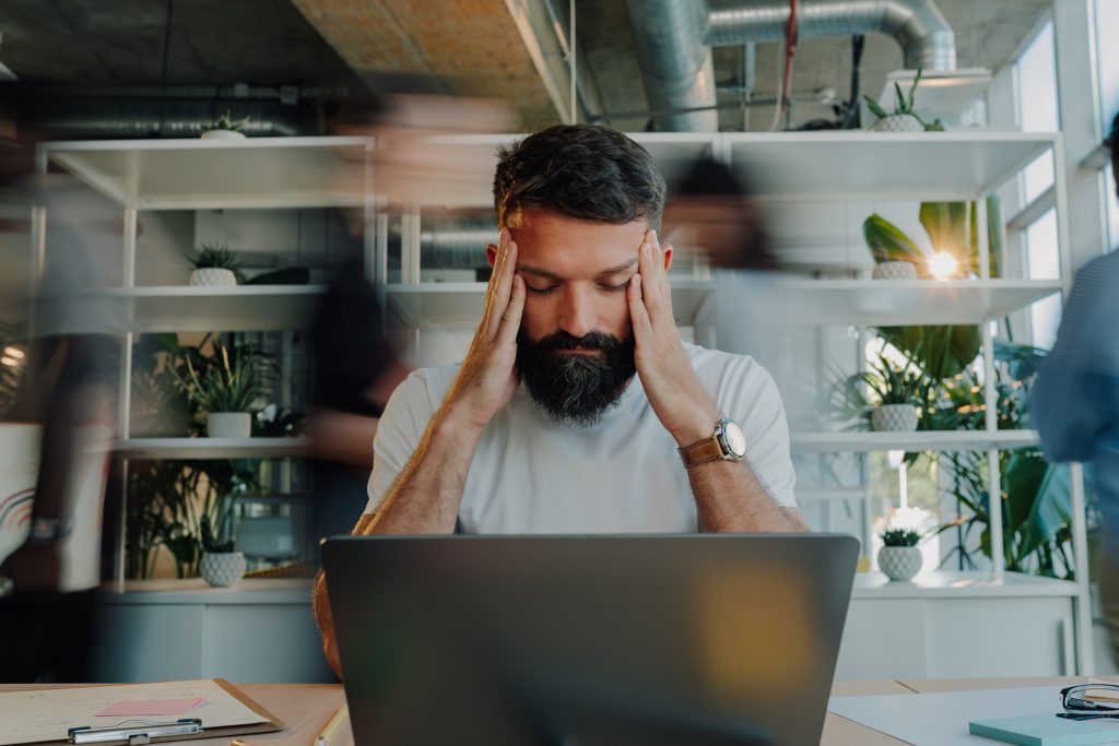 Overworked businessman sitting at his desk, grappling with a headache while managing a laptop in a bustling office environment